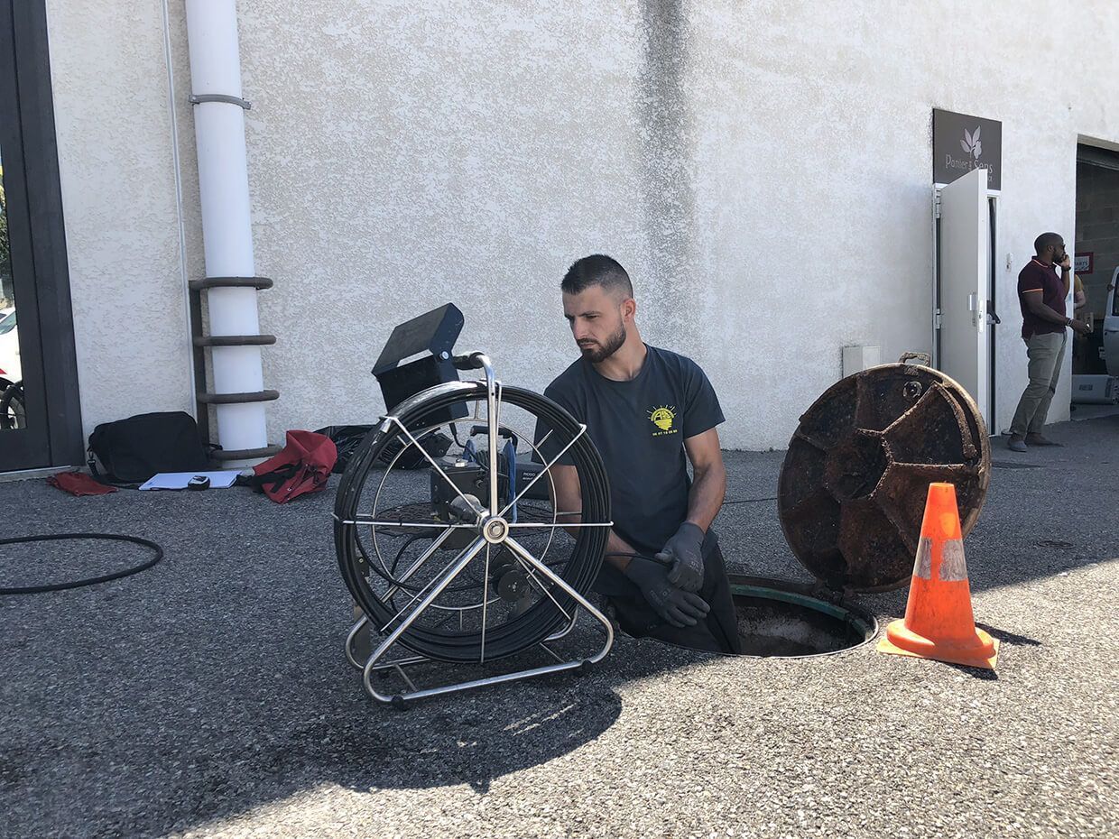Un ouvrier agenouillé près d'un grand ensemble de roues à côté d'un bâtiment, avec un cône de signalisation orange à proximité.