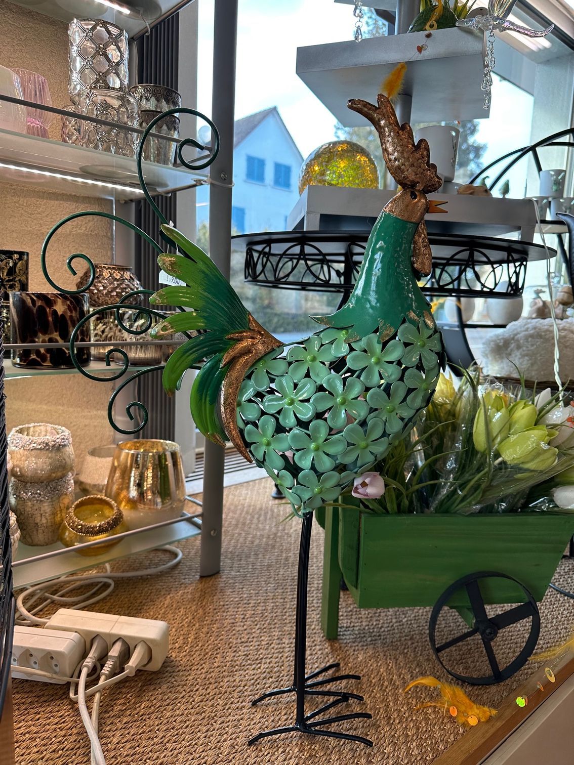 Eine grüne Hahnstatue sitzt auf einem Tisch neben einem grünen Karren voller Blumen.
