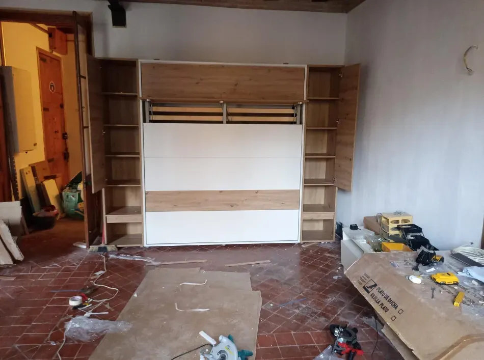 Habitación parcialmente amueblada con un mueble de pared de madera empotrado, suelo de baldosas y herramientas y materiales esparcidos por el espacio.