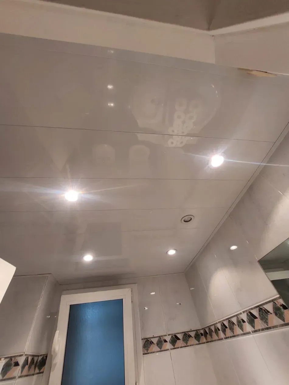 Techo de baño con azulejos blancos brillantes, luces empotradas y una puerta azul debajo.