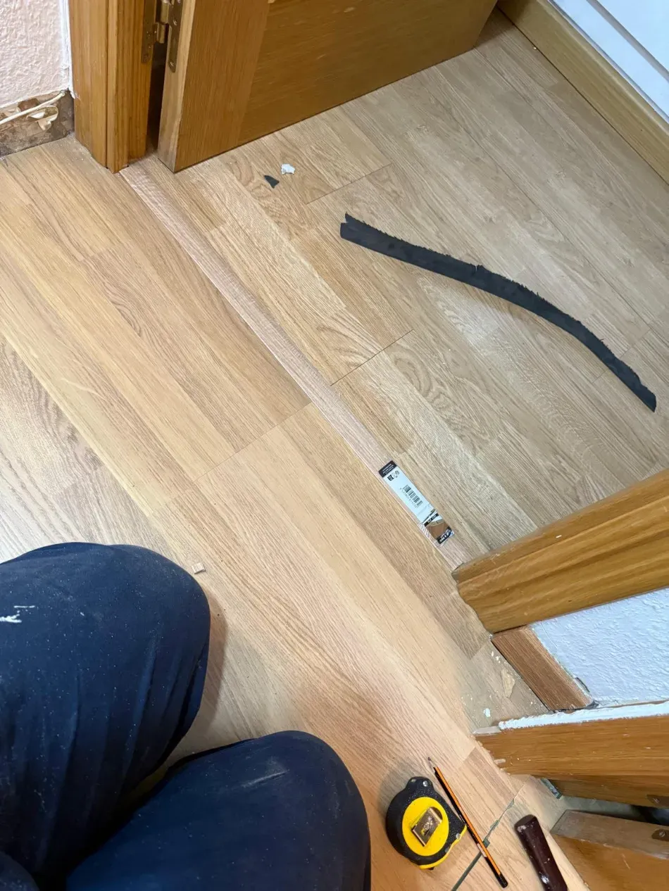 Suelo de madera junto a una puerta con una cinta métrica, una franja negra y la rodilla de una persona visible.