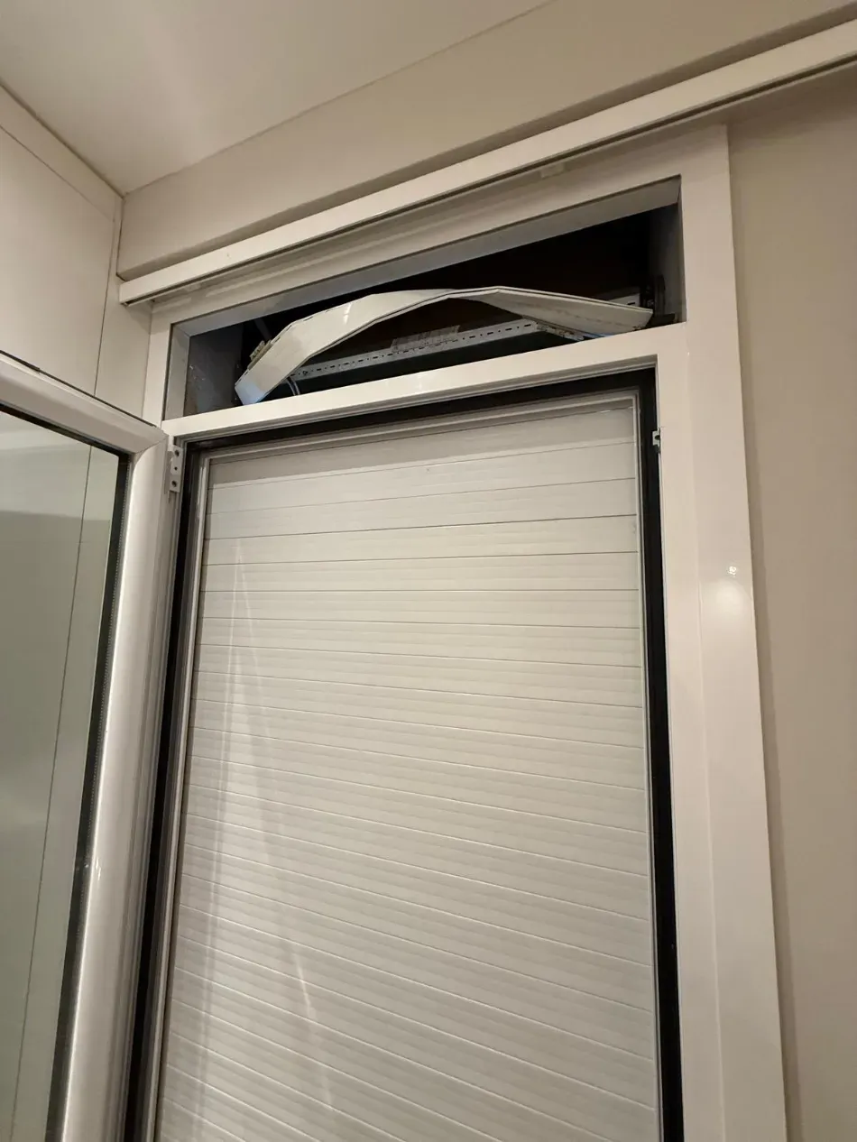 Una persiana enrollable blanca está parcialmente subida en el hueco de una ventana, con una puerta abierta a la izquierda.