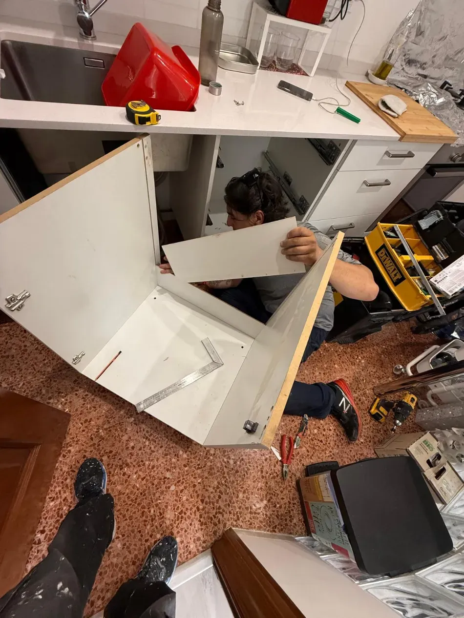 Persona instalando paneles blancos en un armario de la zona de cocina de un taller, con herramientas esparcidas cerca.
