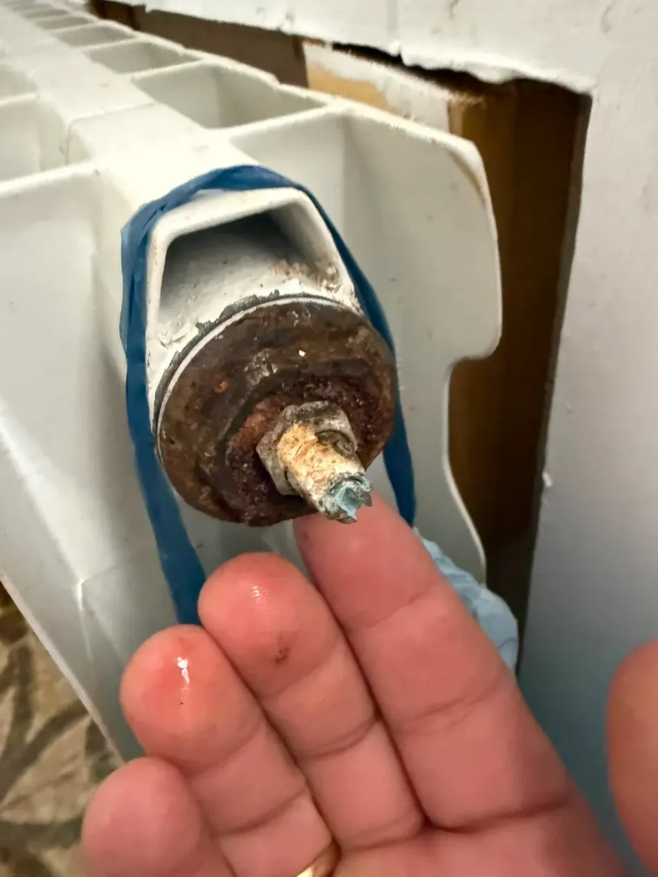 Una mano tocando el vástago de una válvula metálica oxidada en un radiador blanco con cinta azul.