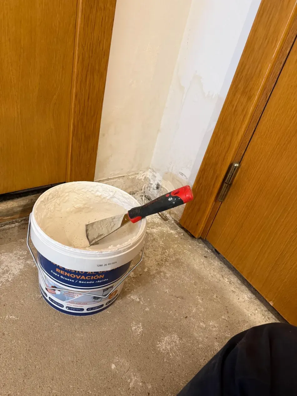 Un cubo de pintura con una espátula en la esquina de una habitación, junto a unas puertas de madera, sobre un polvoriento suelo de hormigón.