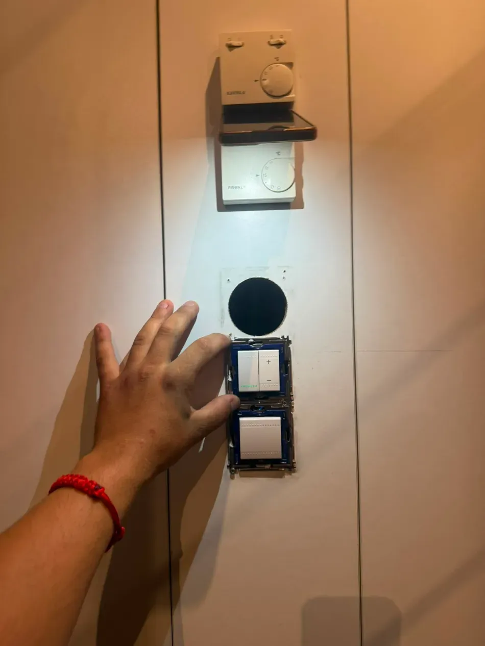 Una mano se extiende hacia un panel de pared con un agujero circular y dos botones cuadrados bajo una luz cálida.