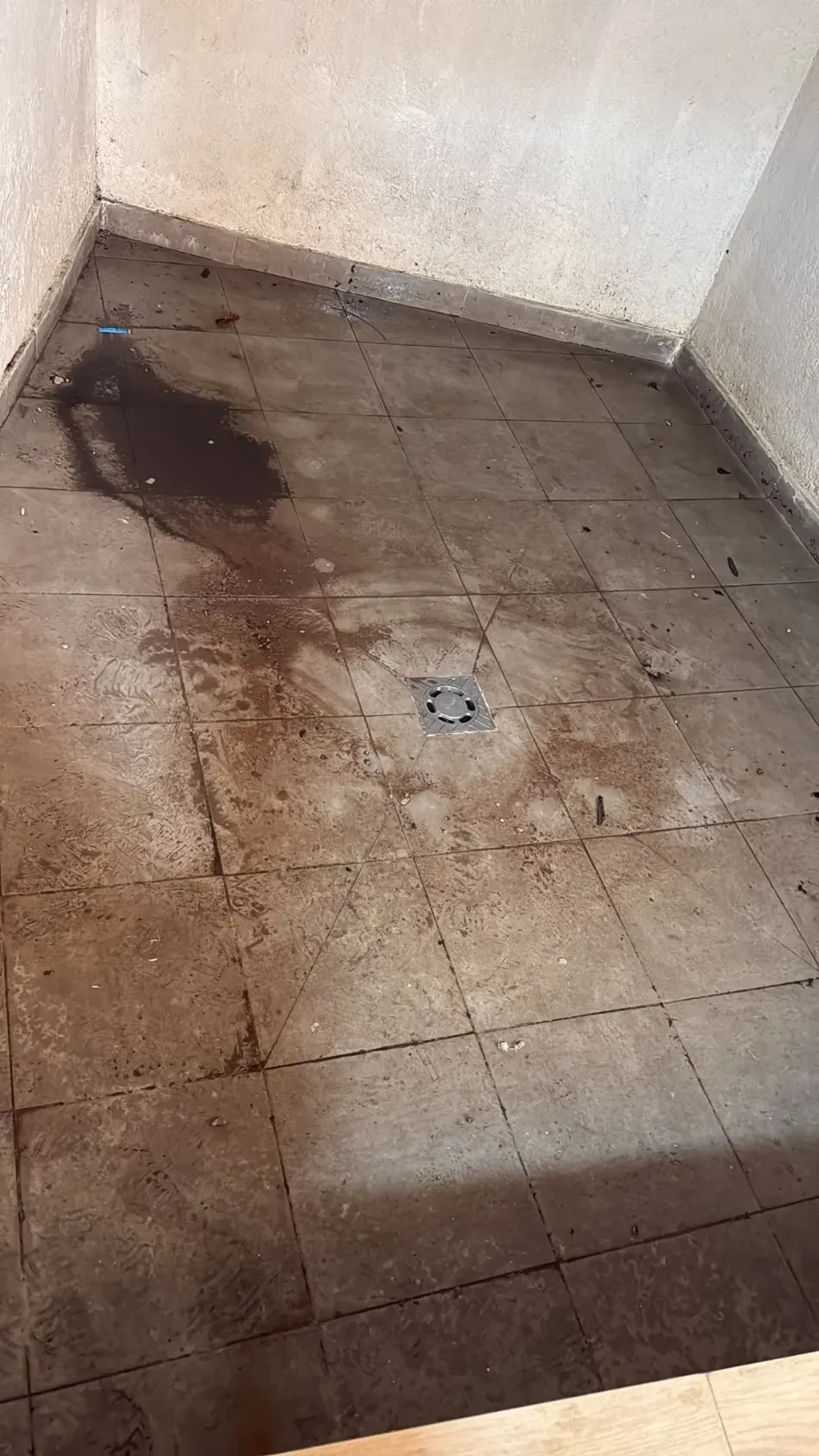 Suelo de baldosas sucio con manchas marrones y un pequeño desagüe en un rincón vacío de una habitación.