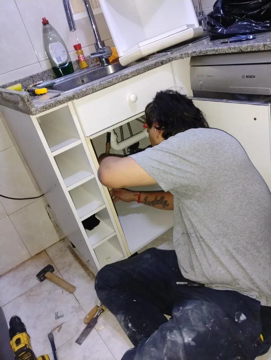 Una persona repara un mueble bajo el fregadero con herramientas en el suelo de la cocina.