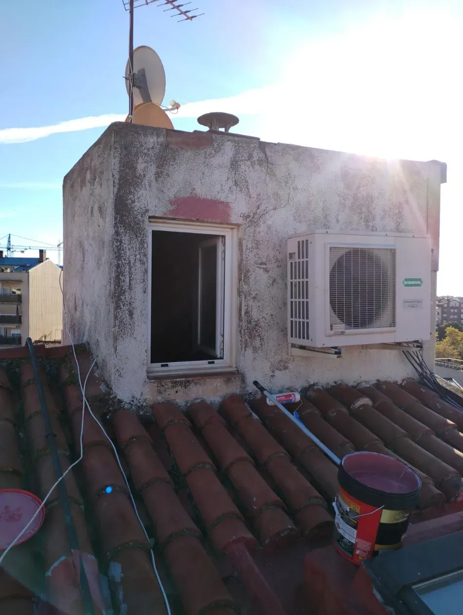 Azotea con edificio blanco desgastado, ventana abierta, aire acondicionado, antena parabólica y luz solar brillante.