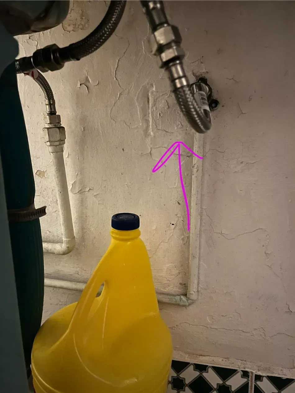 Jarra amarilla debajo de tuberías expuestas contra una pared, con una flecha rosa que apunta a una conexión de tubería.