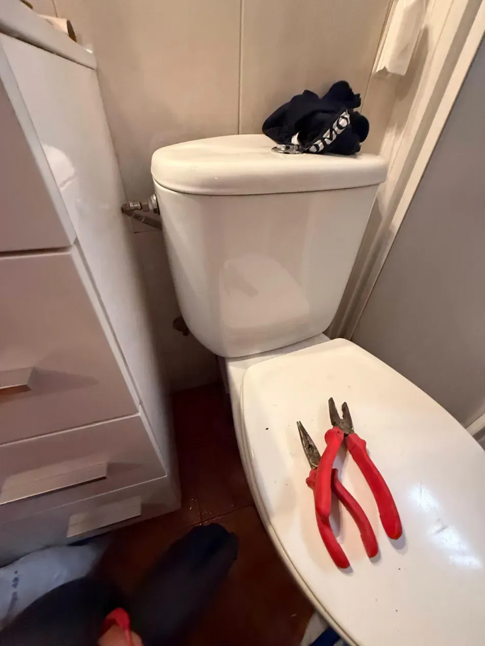 Baño pequeño con inodoro, alicates de mango rojo sobre la tapa y zapatos en el suelo.