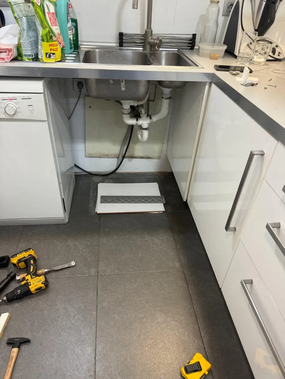 Área del fregadero de la cocina con tuberías expuestas, productos de limpieza y herramientas en el suelo durante las reparaciones.