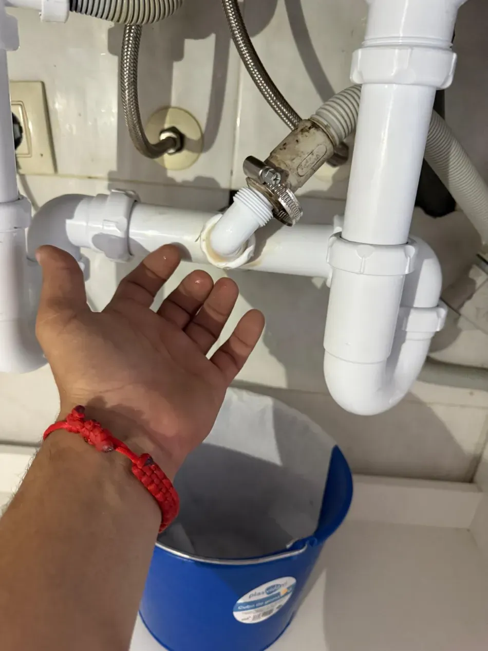 Una mano se introduce debajo de un fregadero para inspeccionar las tuberías de PVC blancas, con un cubo azul debajo.