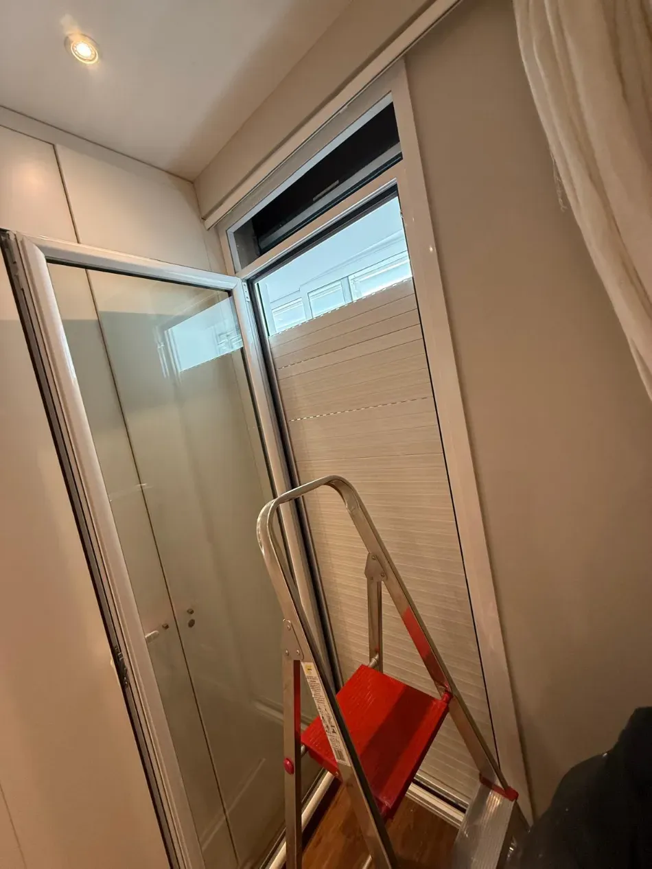 Baño con puerta de ducha de cristal, persiana y escalera plateada con bandeja roja.