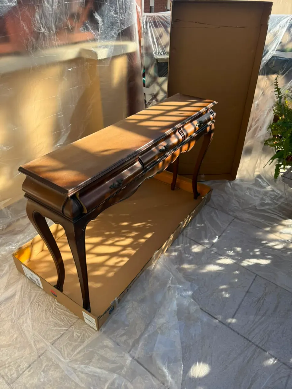 Banco de madera en un patio soleado al aire libre, con respaldo marrón y patas de metal sobre un suelo cubierto de plástico.