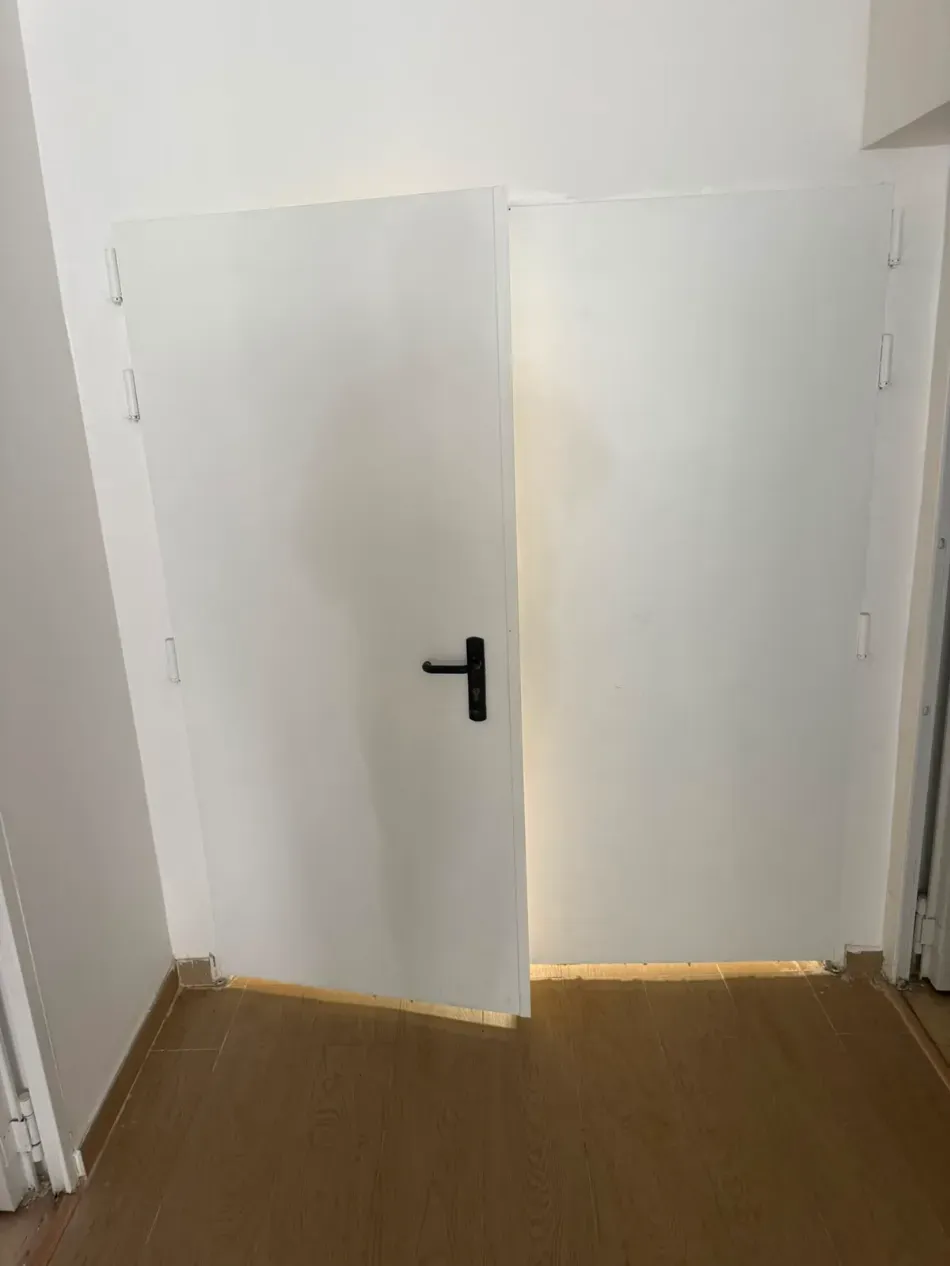En un pasillo estrecho con suelo de madera, unas puertas dobles blancas, ligeramente abiertas y con tirador negro, se encuentran.