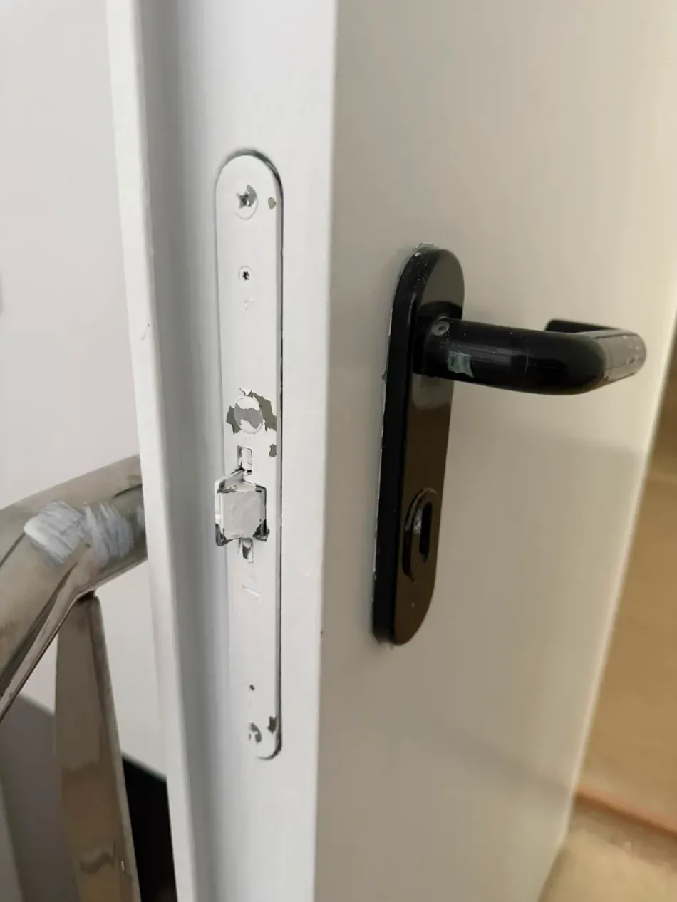 Primer plano de una puerta blanca con manija negra y placa de cerradura dañada.