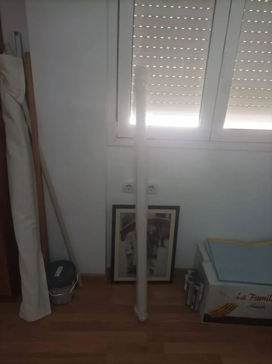 Rincón del dormitorio con dos barras blancas, cortinas enrolladas, cuadros enmarcados y una caja junto a una ventana con contraventanas.