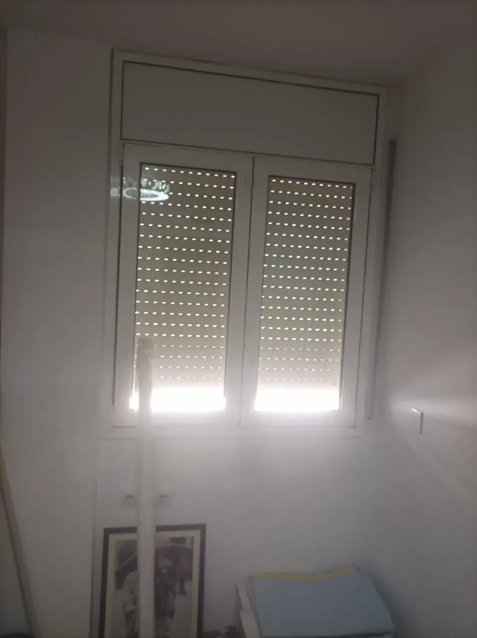 Ventana soleada con persianas parcialmente cerradas en una habitación sencilla, con fotos enmarcadas y una pequeña mesa debajo.