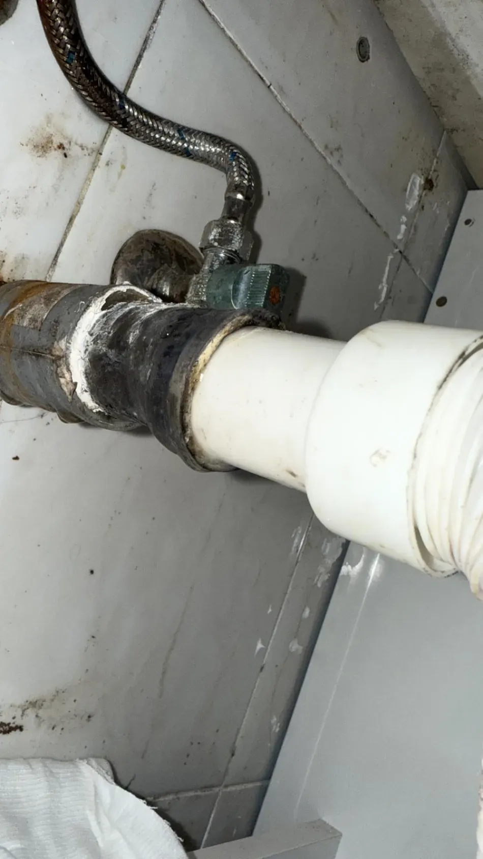 Primer plano de las conexiones corroídas de una tubería y de una tubería de PVC blanca contra una pared gris.