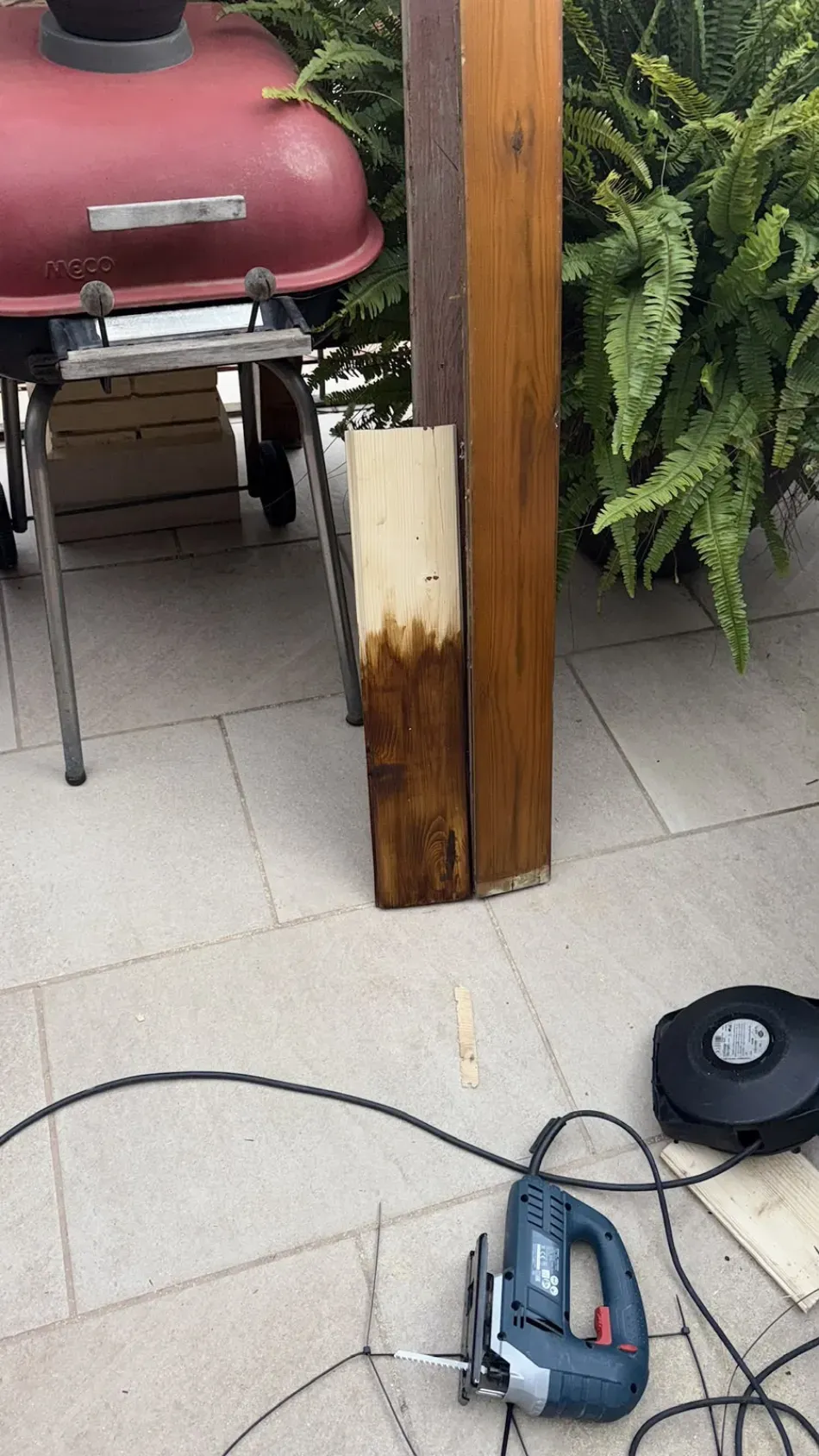 Un poste de madera carbonizado junto a una parrilla en un patio embaldosado, con una herramienta eléctrica y un cable en el suelo.