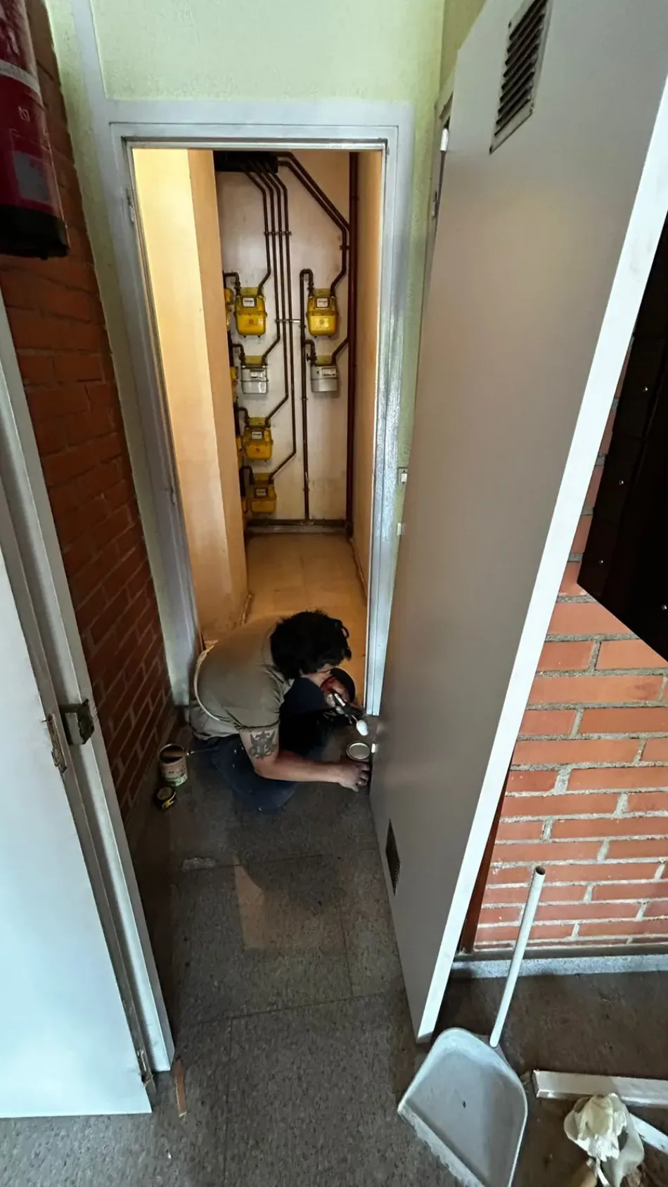 Una persona se encuentra agachada en un pasillo estrecho cerca de una puerta, con una pared de ladrillos y contadores de servicios públicos a la vista.