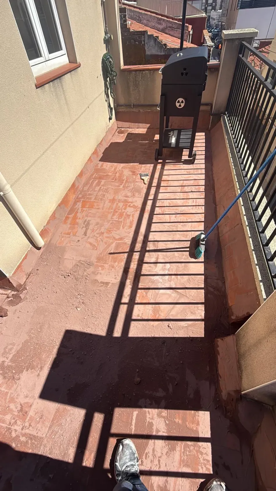 Balcón soleado de un apartamento con reja negra, pasillo estrecho y fuertes barandillas que proyectan sombras en el suelo.
