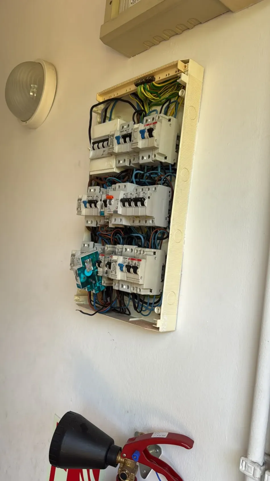 Panel de interruptores eléctricos abierto empotrado en la pared, con una lámpara de pared negra y una herramienta roja debajo.