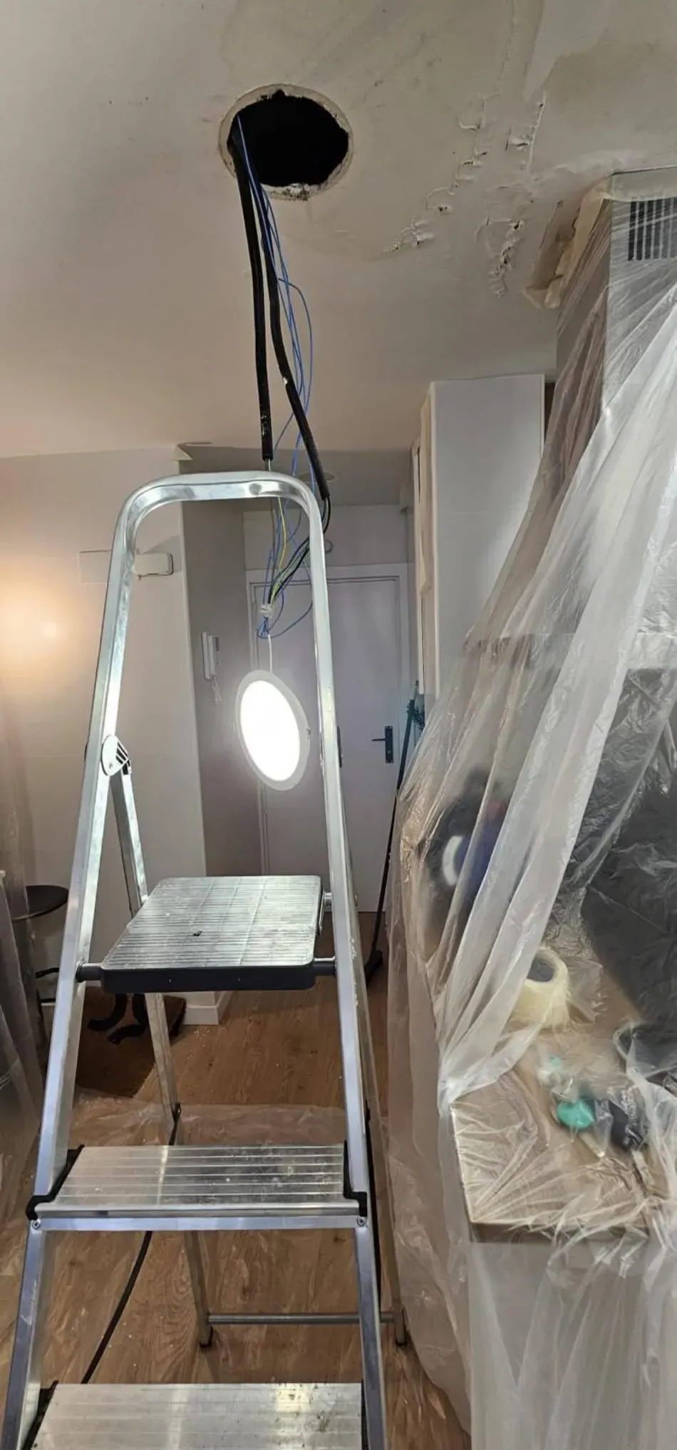 Escalera de mano en una habitación en obras, con una lámina de plástico y una abertura para una lámpara de techo colgante.