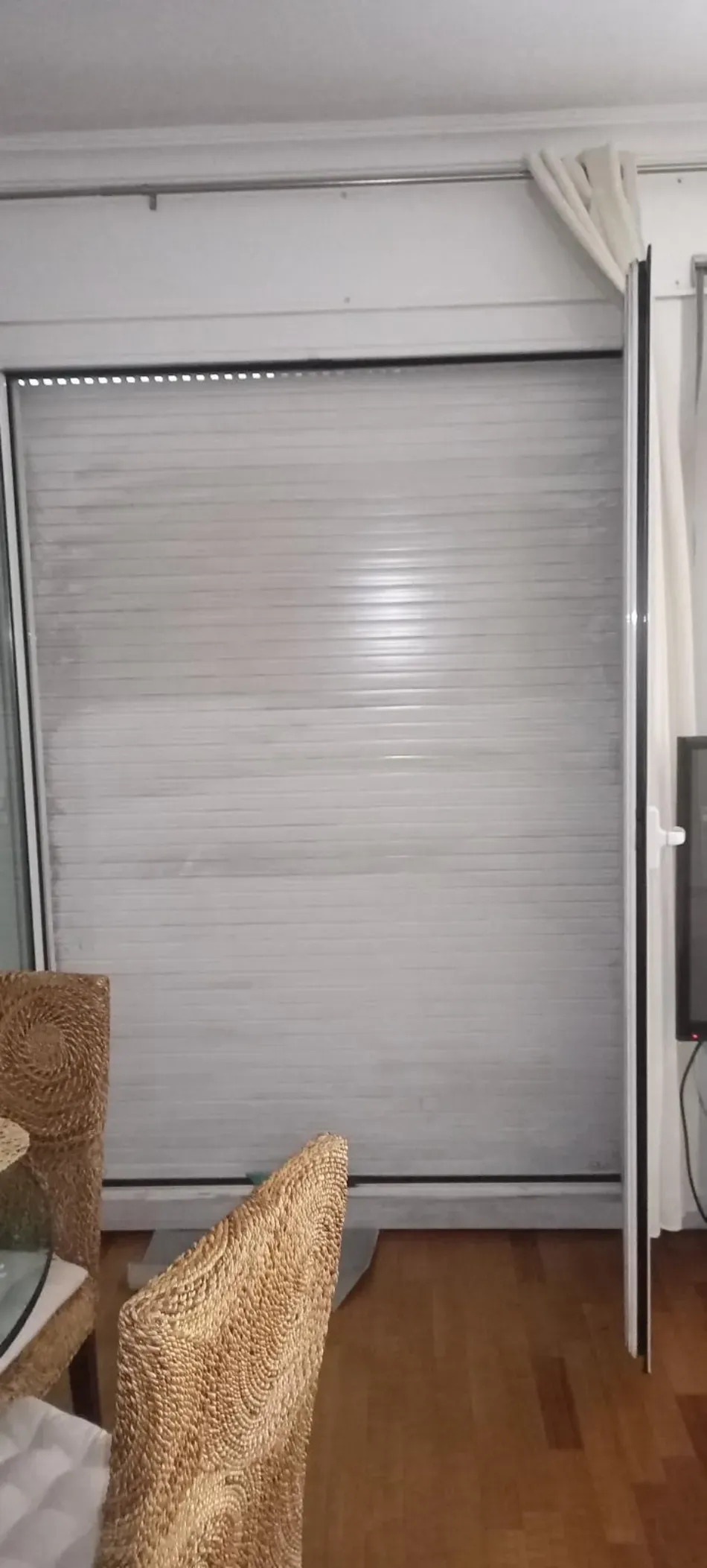 Estor enrollable gris cerrado que cubre una ventana en una sala de estar con piso de madera y borde de sofá.