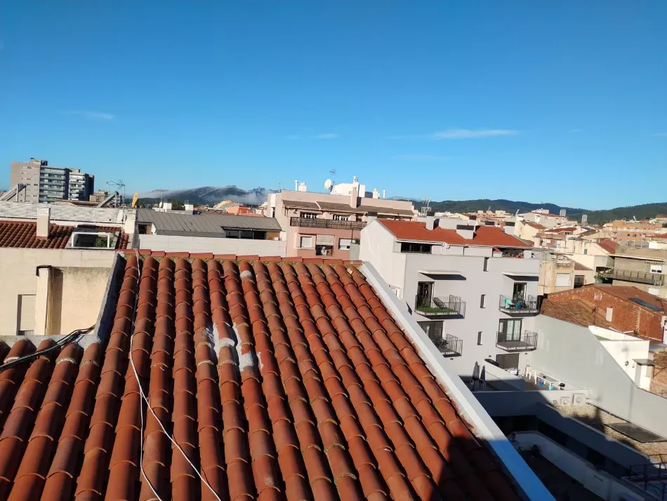 Vista desde la azotea de tejas de terracota con vistas a una ciudad iluminada por el sol y colinas distantes bajo un cielo azul despejado.