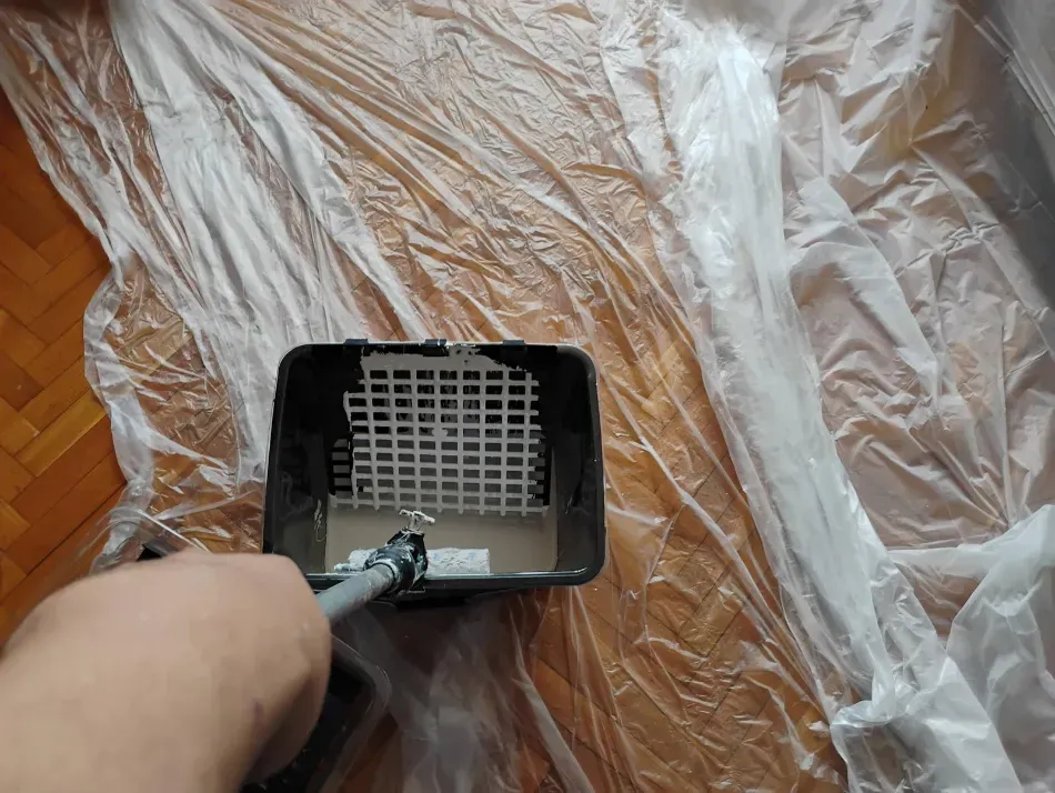 Mano sosteniendo un rodillo de pintura sobre una bandeja negra en un suelo de madera cubierto de plástico.