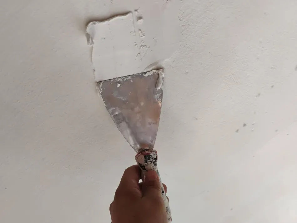Mano sosteniendo una espátula, raspando pintura blanca de un parche en la pared.