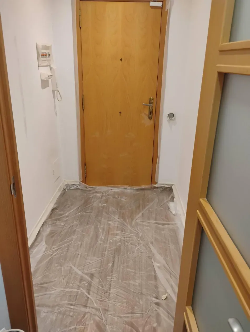 Pasillo estrecho con puerta de madera, paredes blancas y suelo cubierto de plástico en obras.