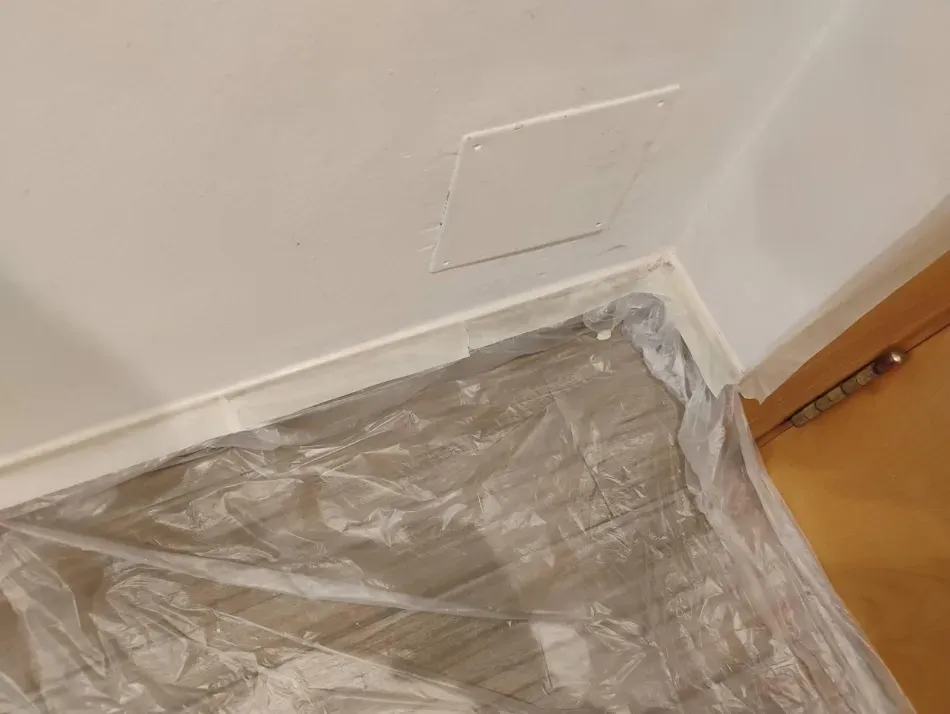 Suelo cubierto de plástico en la esquina de una habitación junto a una pared blanca y un marco de puerta de madera.