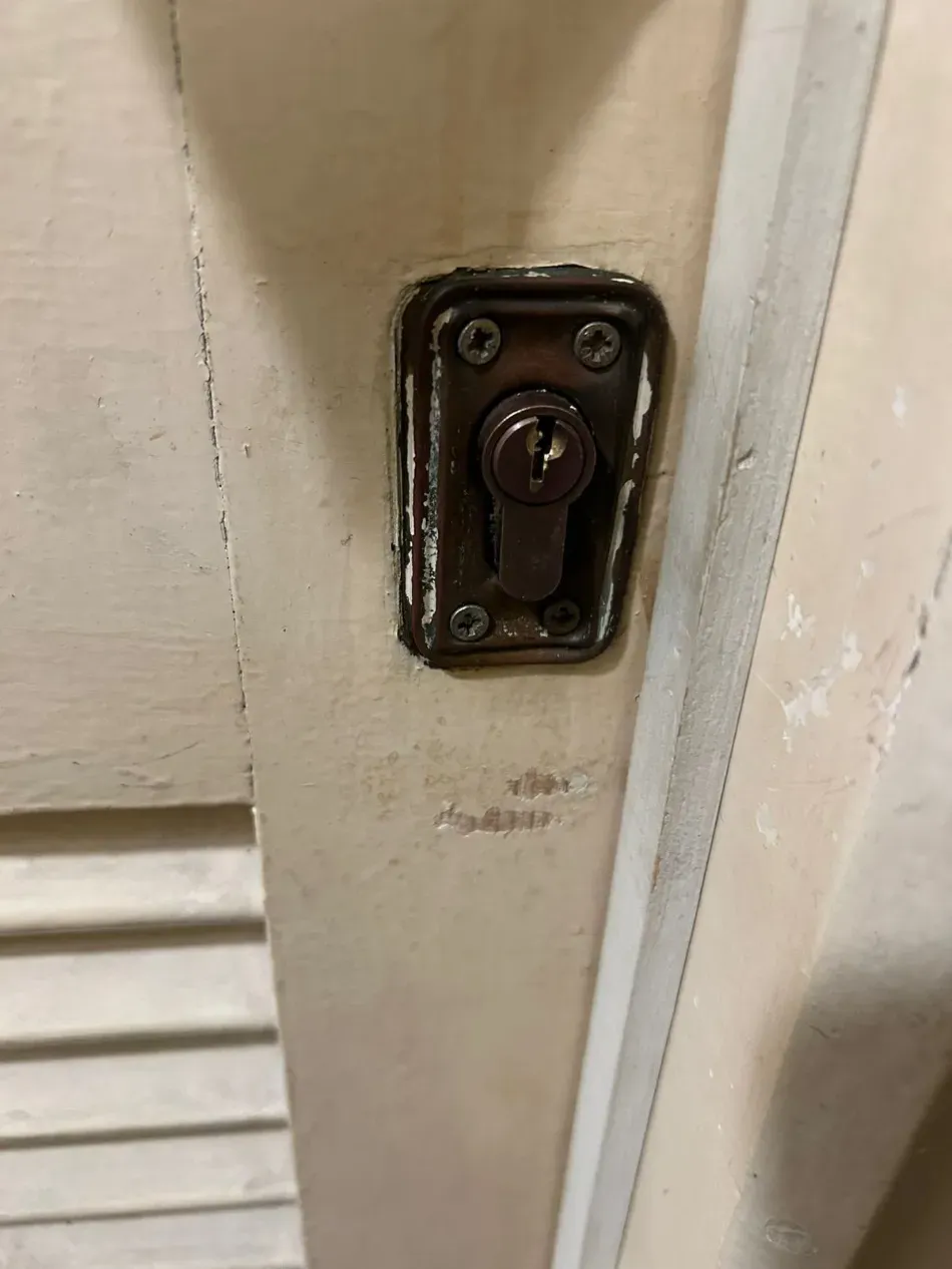 Primer plano de un pestillo metálico desgastado en un marco de puerta de madera pintado de color crema.