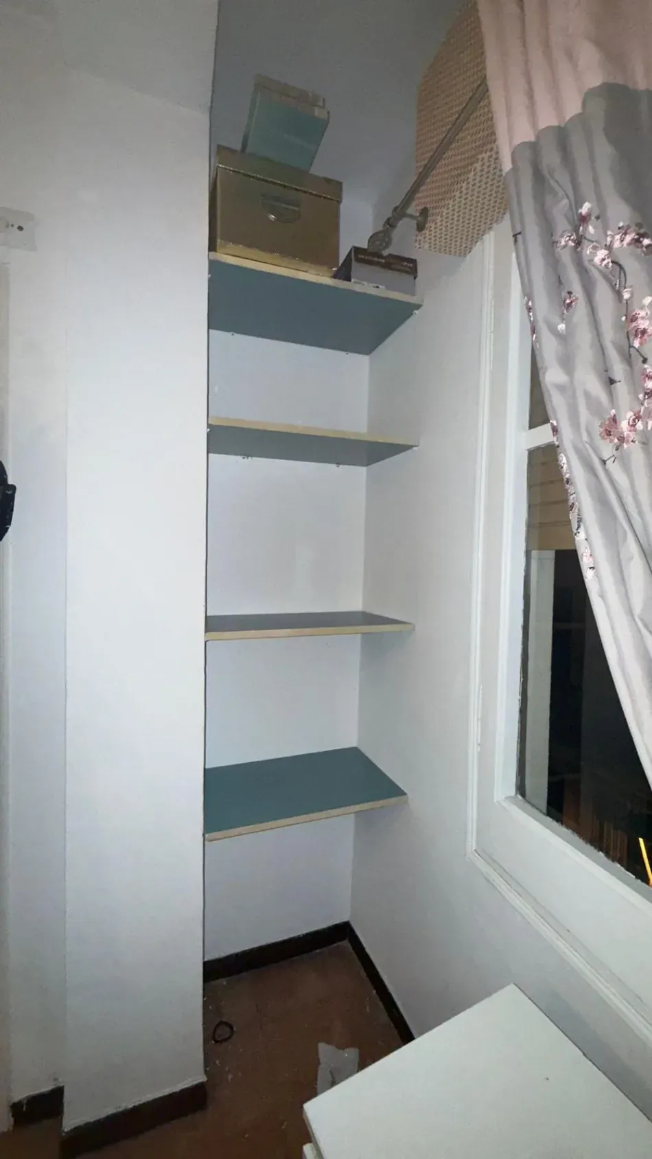 Estantería alta de esquina con estantes azules junto a una ventana y una cortina en una habitación pequeña.