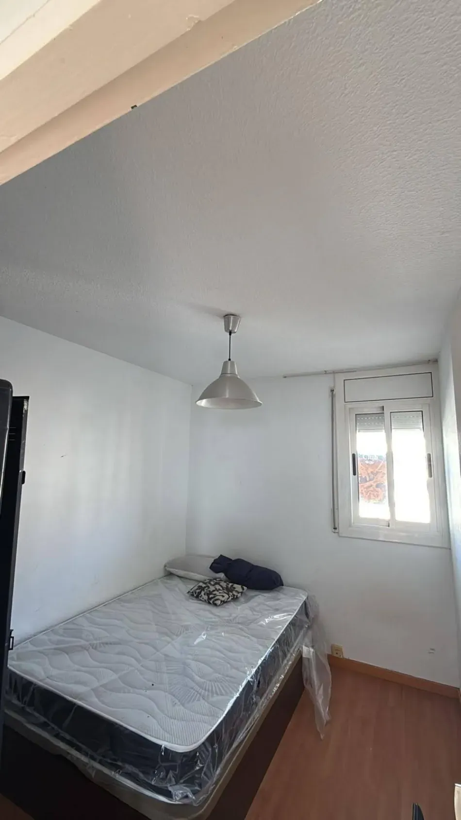 Habitación pequeña con una cama individual, paredes blancas, suelo de madera y una lámpara de techo colgante.