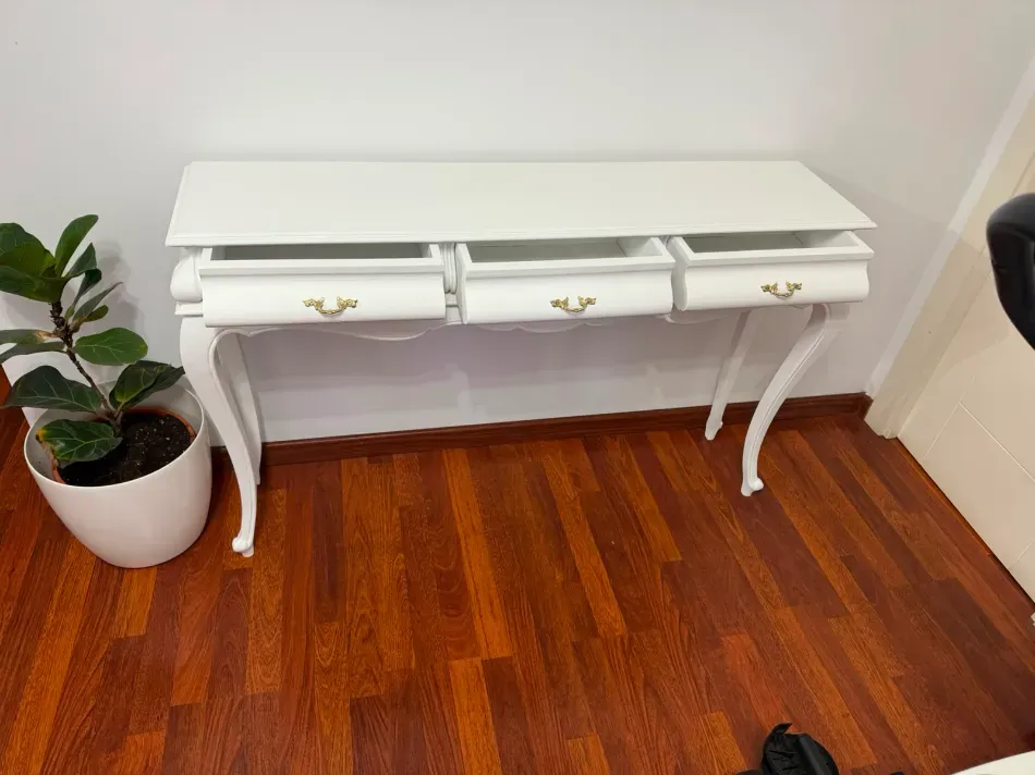 Consola blanca con tres cajones contra la pared, junto a una planta en maceta sobre un suelo de madera.