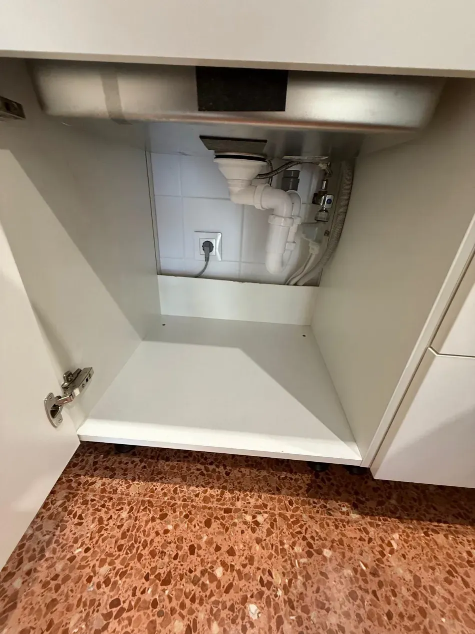 Mueble bajo el fregadero con tuberías a la vista e interior blanco, abierto sobre un suelo moteado de color marrón.