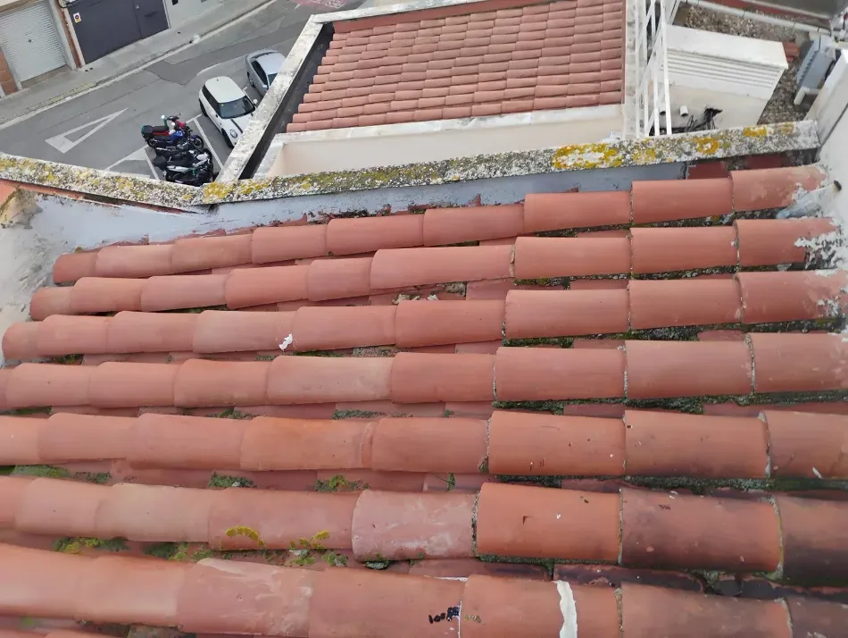 Tejado de tejas de arcilla roja con una terraza adyacente, barandilla, motos aparcadas y edificios de ladrillo al fondo.