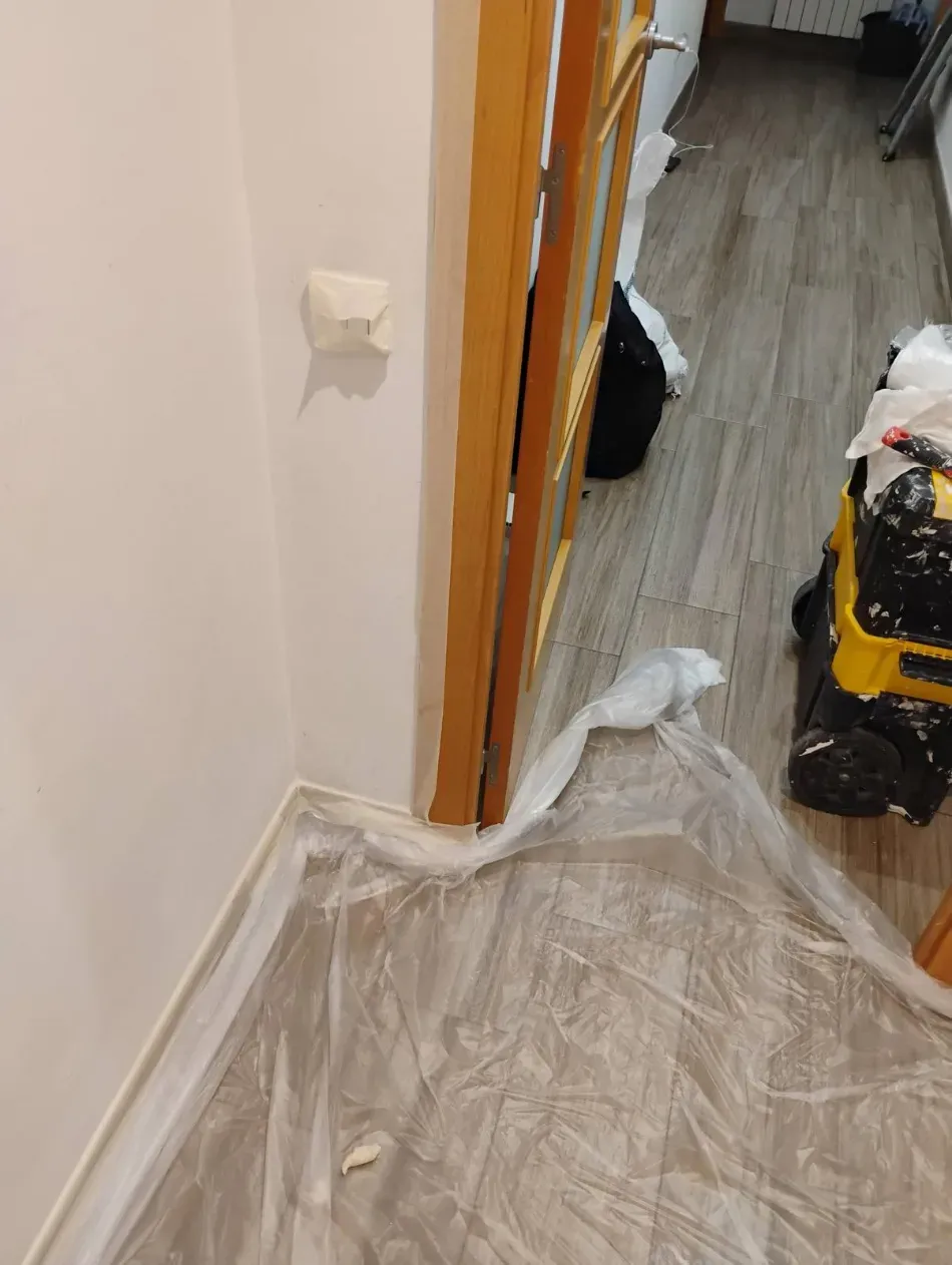 Pasillo estrecho con el suelo cubierto con una lámina de plástico y una puerta que da a una habitación con productos de limpieza.