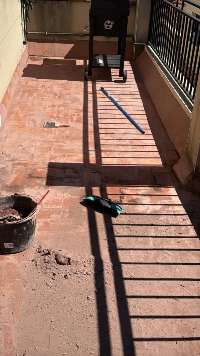 Balcón de terracota bañado por el sol, con barandilla negra, sombras y una escoba en el suelo.