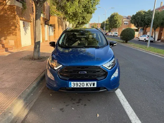 Ford EcoSport azul estacionado en una calle con un edificio de ladrillo y árboles al fondo.