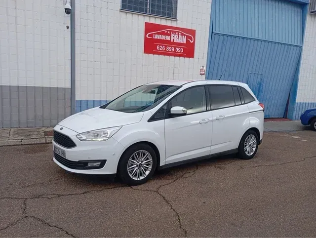 Un Ford C-Max blanco estacionado frente a un edificio azul y blanco, con un cartel comercial en la parte superior.