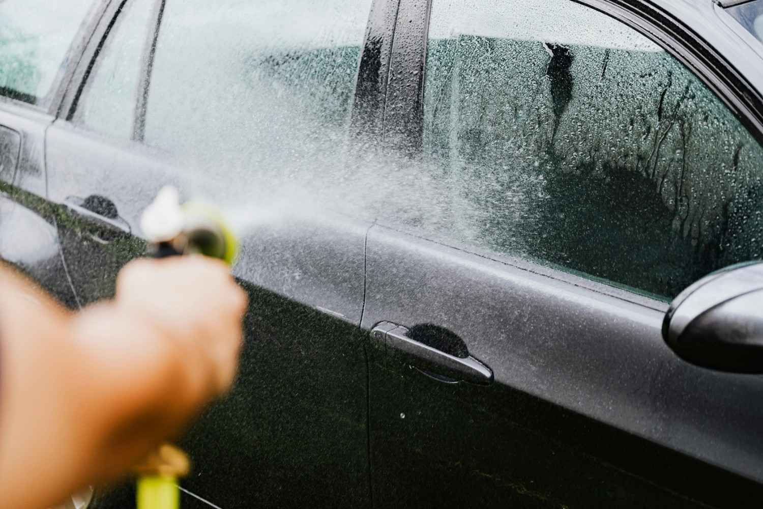 Persona rociando agua sobre un automóvil negro con una manguera.