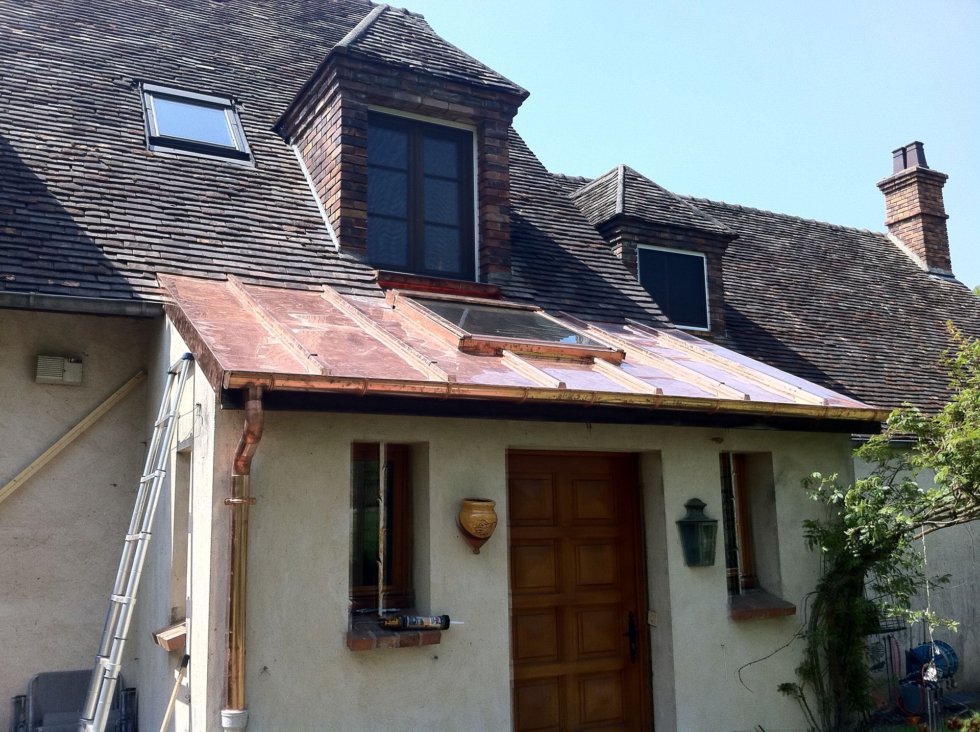 Entrée d'une vieille maison rustique avec toit de tuiles, porte en bois et lucarnes sous des bardeaux usés par le temps.