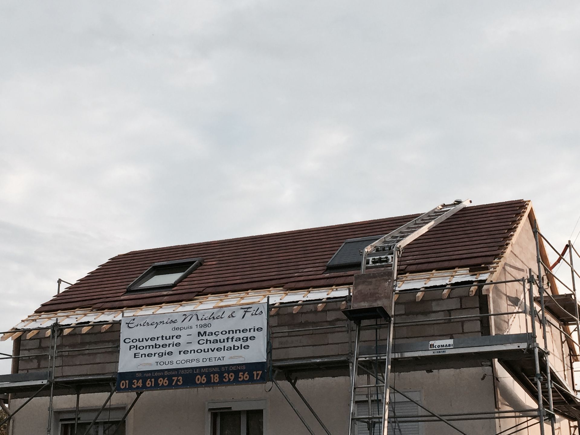 Toiture d'une maison partiellement réparée, avec échafaudages et grande banderole sous un ciel nuageux.