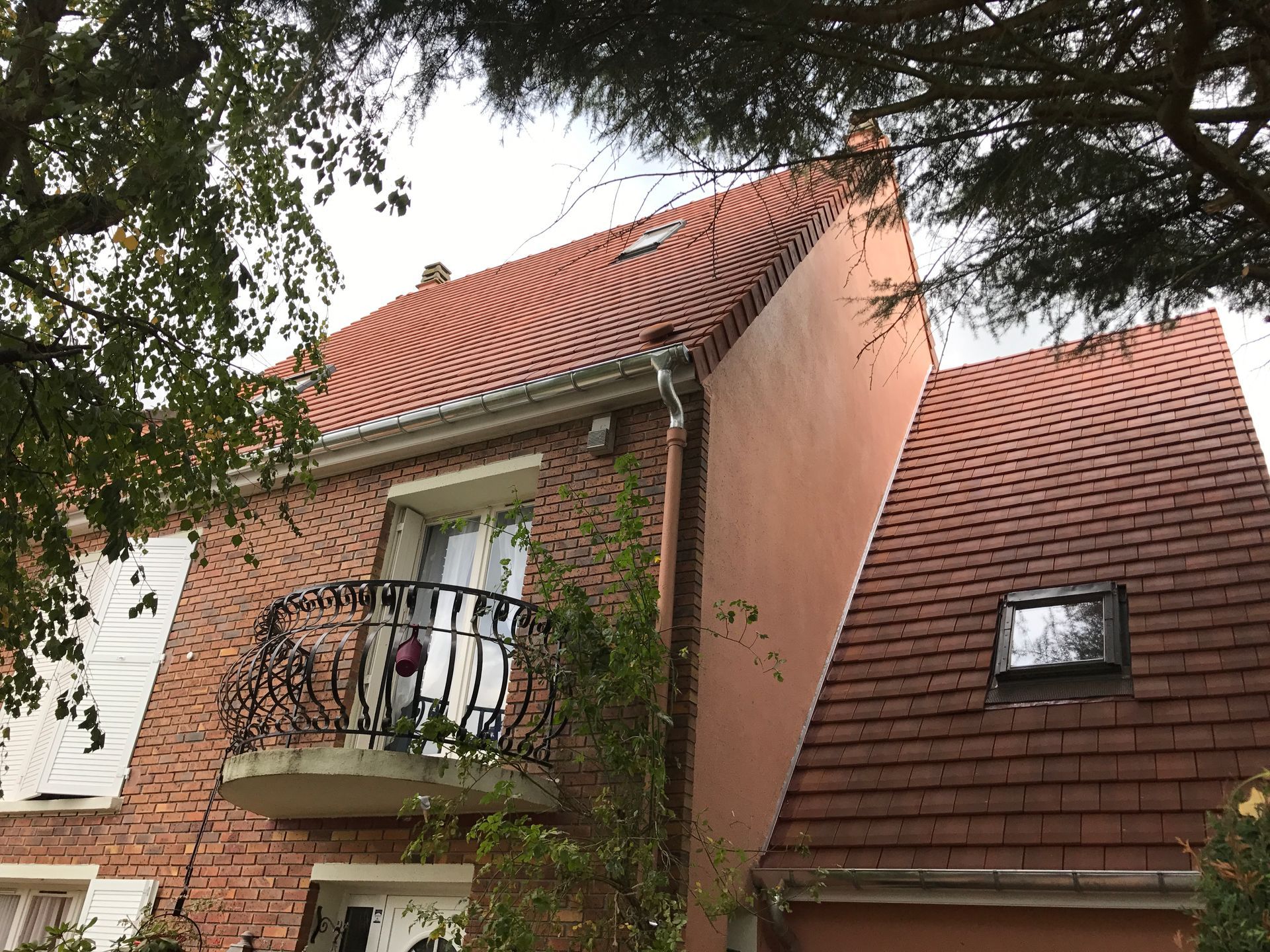 Maison en briques avec toit de tuiles rouges et petit balcon, entourée d'arbres.