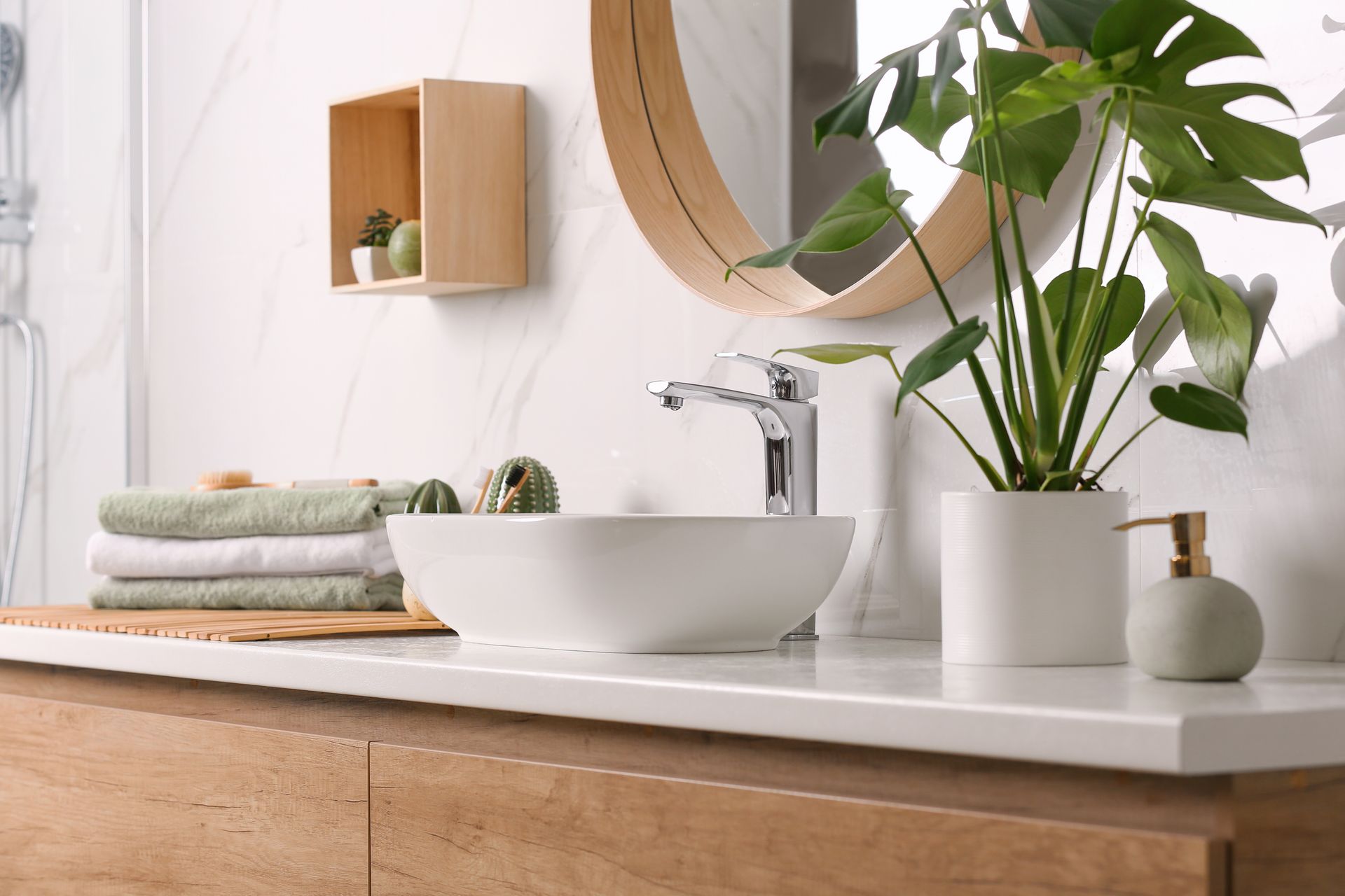 Meuble sous lavabo moderne avec lavabo blanc, robinet chromé, miroir rond et plante en pot à côté de serviettes pliées.